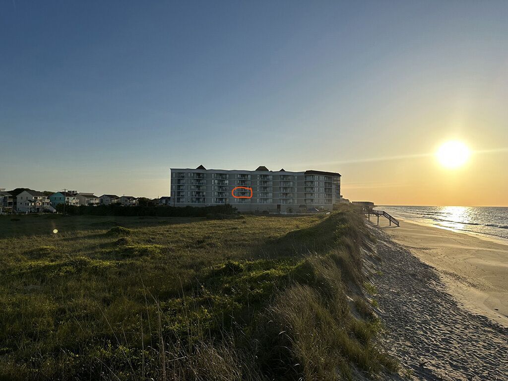 A beachfront hotel with a long pier extending into the ocean, surrounded by grassy dunes and other buildings.