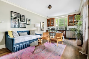 A living room with a blue sofa, a coffee table, and a rug. There are framed pictures on the wall, a lamp, and a kitchen in the background.
