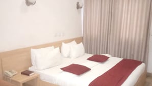 Une chambre d’hôtel avec un lit en bois, du linge de lit blanc, un plaid rouge, une table de chevet avec un téléphone et un livre, et un appareil de climatisation fixé au mur.