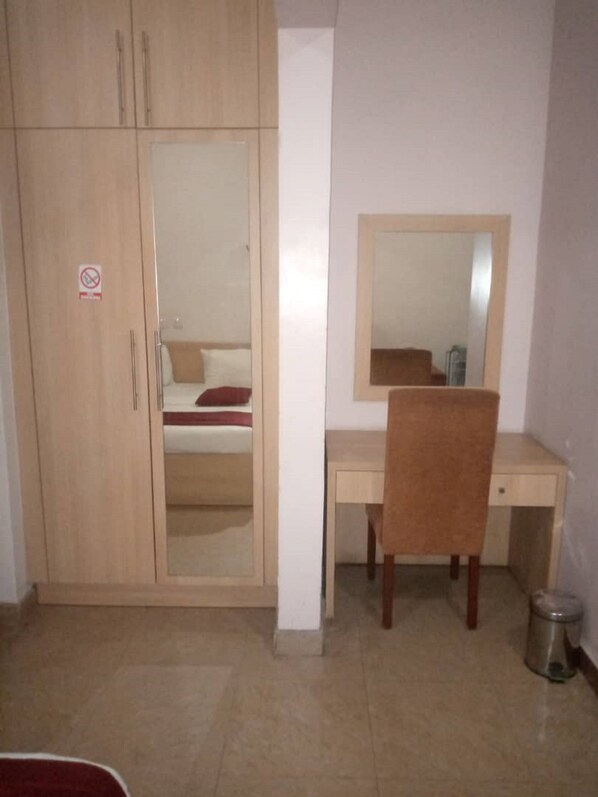 A bedroom with a wooden wardrobe, a desk with a chair, a mirror, and a bed with a red pillow.