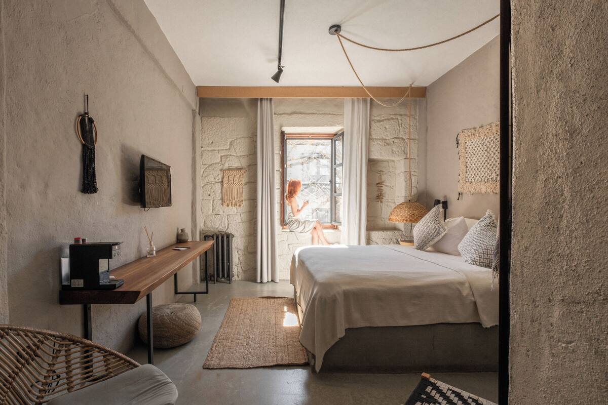Un dormitorio con cama, una mesita de noche de madera, una silla, una lámpara y vistas al exterior.