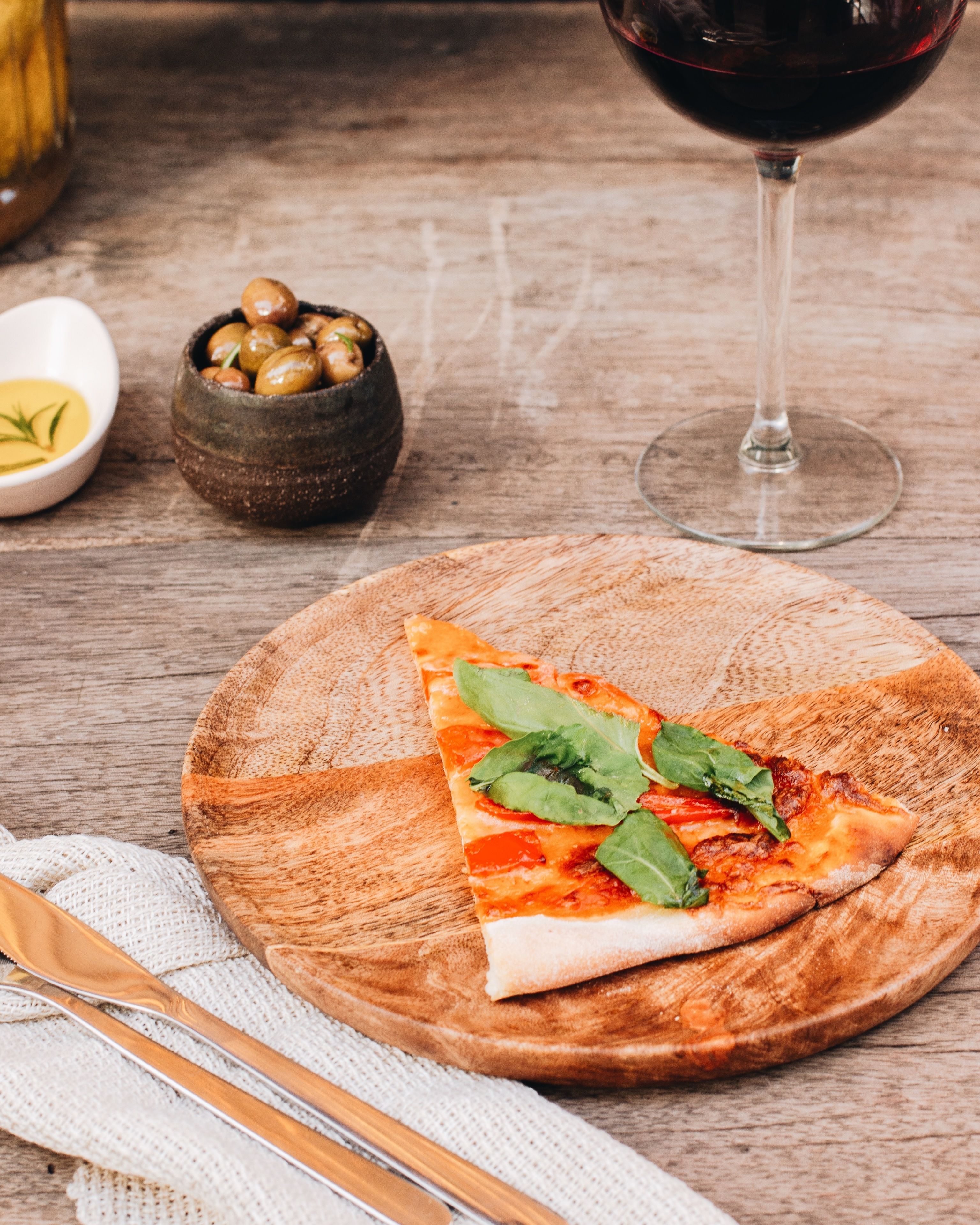 A slice of pizza on a wooden plate with a glass of red wine and a bowl of olives in the background.