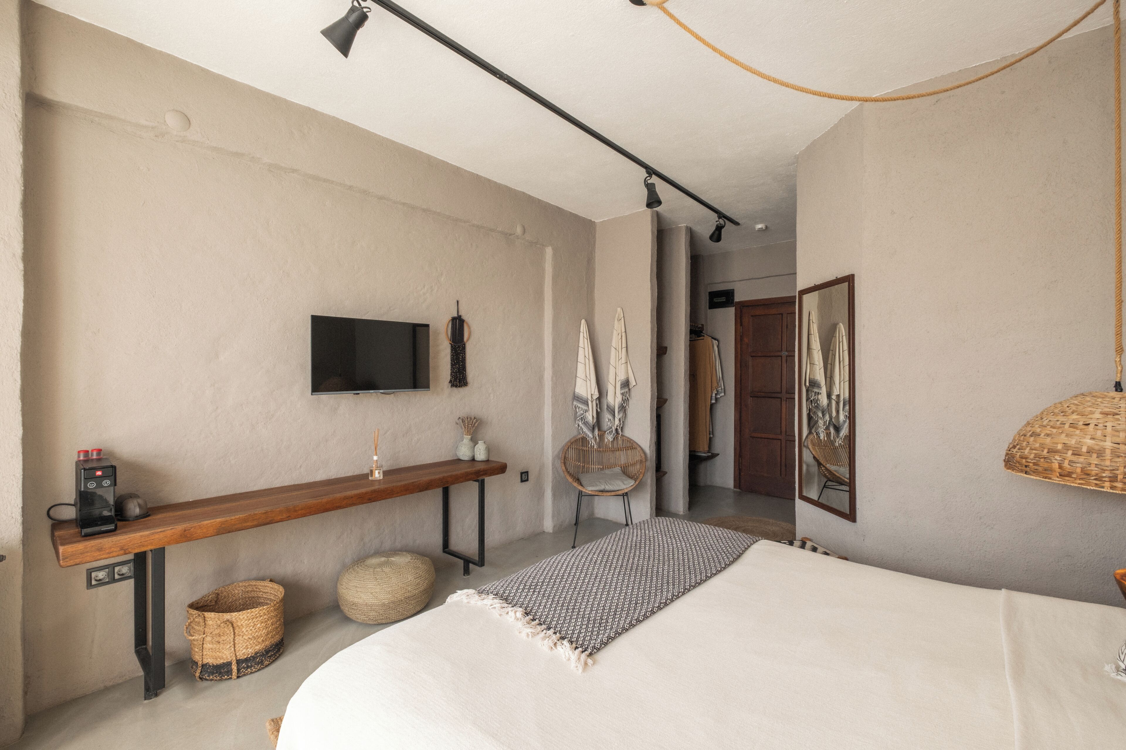 Un dormitorio con cama, televisor montado en la pared, banco de madera, silla de mimbre y lámpara colgante.