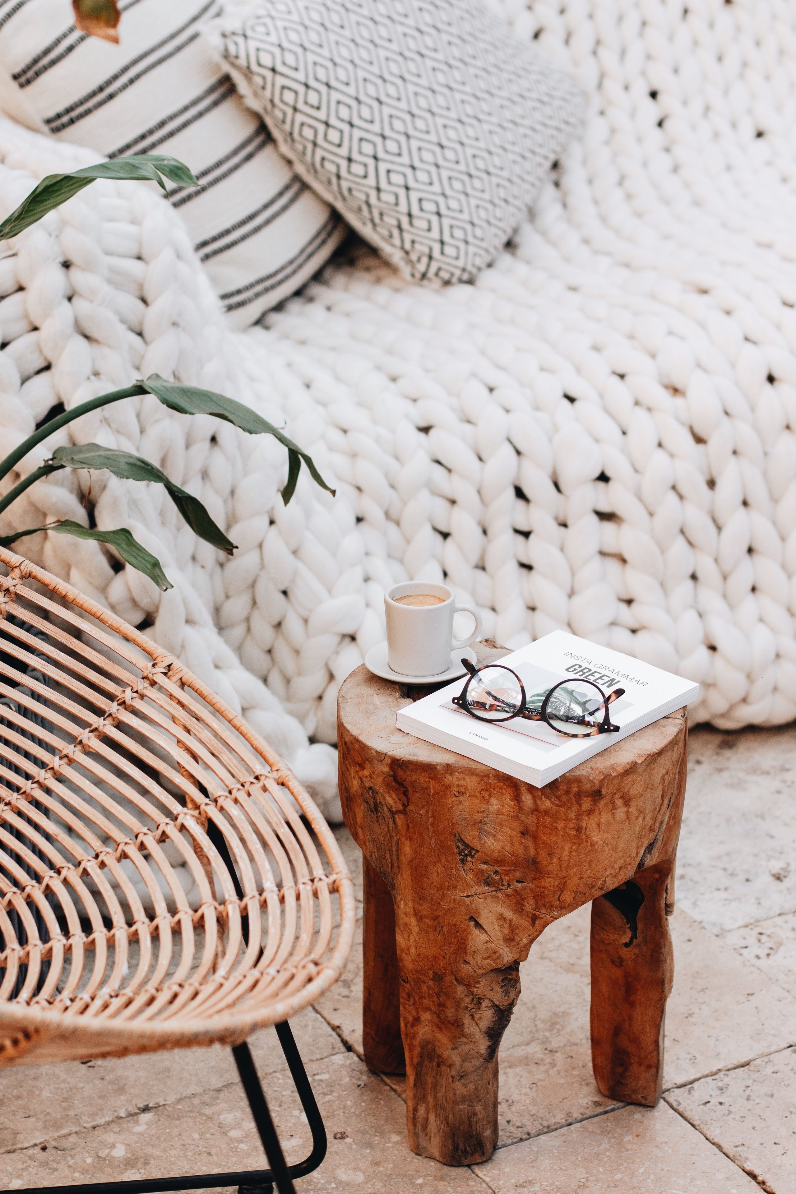 A wicker chair, a small wooden stool, a book, and a cup of coffee on a woven blanket.