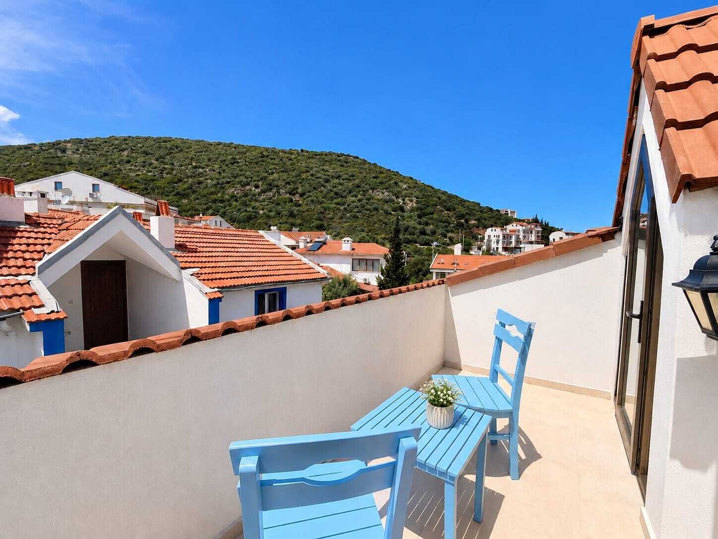 A balcony with a table and chairs, a potted plant, and a view of a hillside with buildings.