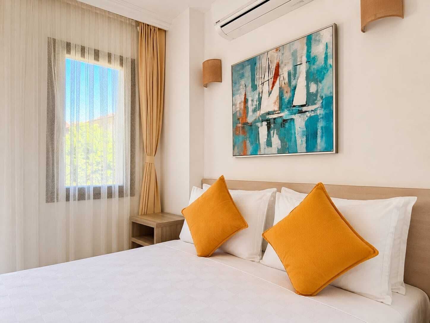Habitación de hotel con cama, dos almohadas, mesita de noche, un cuadro y ventana con cortinas.