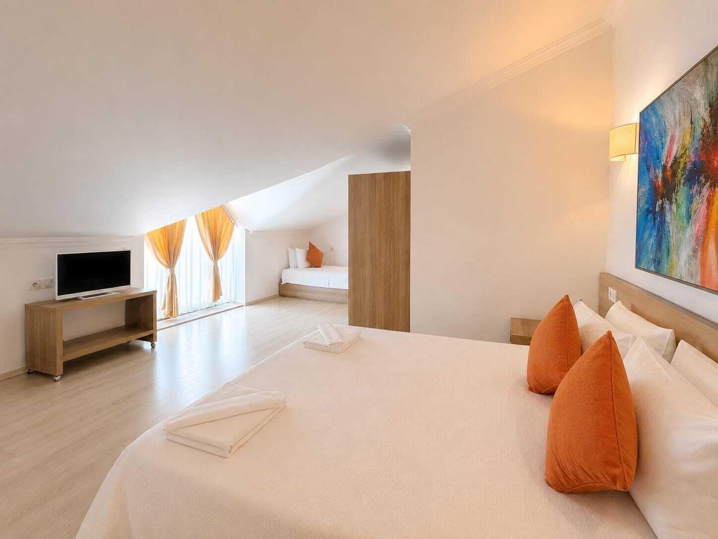 A hotel room with a large bed, a TV on a wooden stand, a painting on the wall, and a window with curtains.