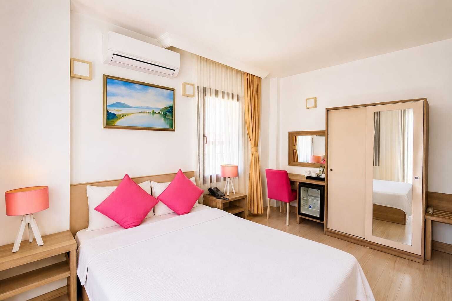 A hotel room with a bed, bedside lamps, a desk, a mirror, and an air conditioning unit.