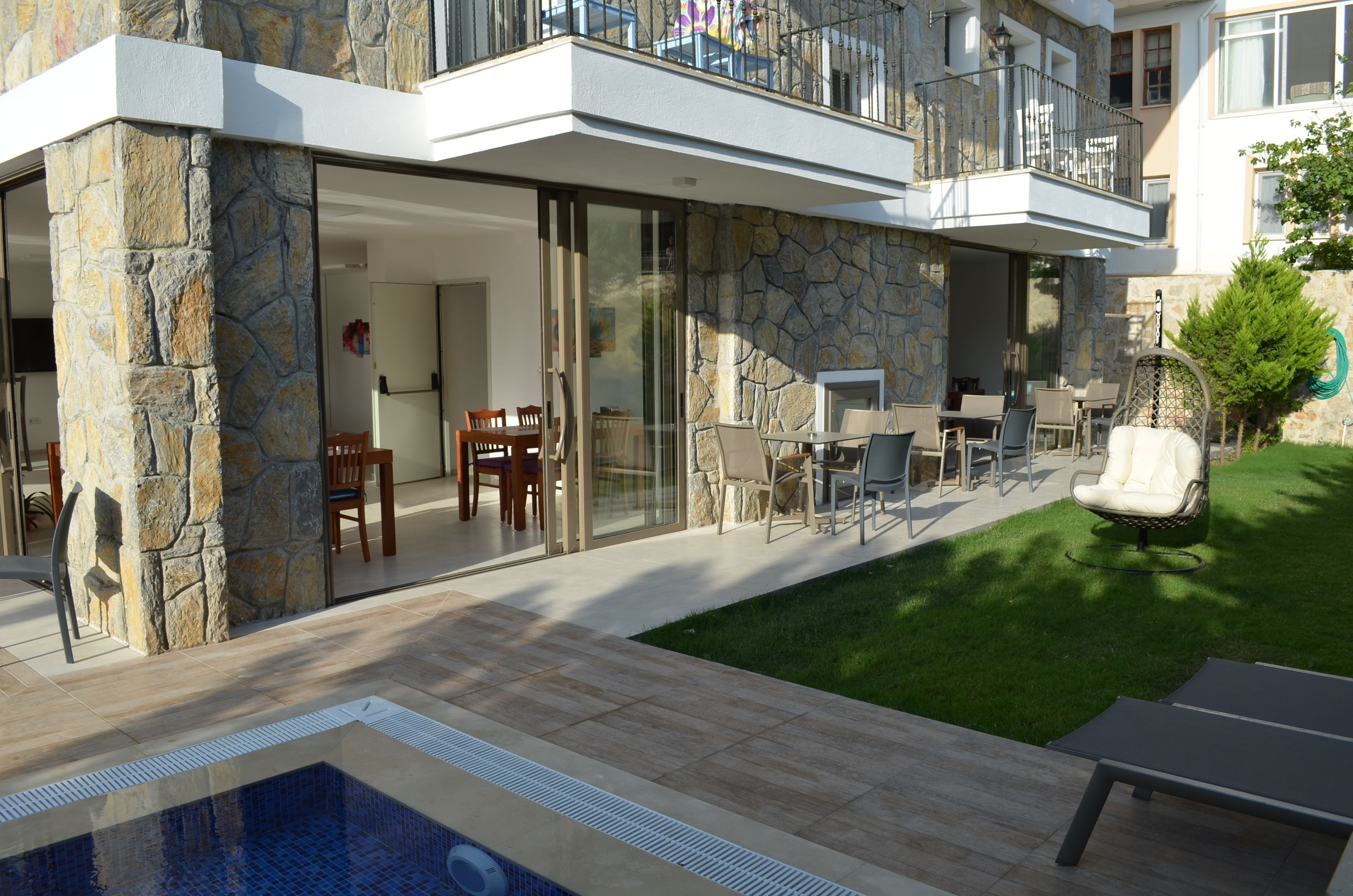 Une maison moderne avec une façade en pierre, une porte en verre donnant sur un espace repas extérieur et une piscine.