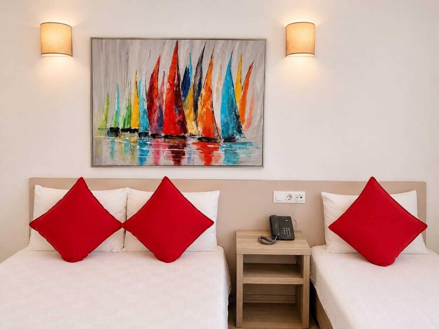 A hotel room with two beds, each with a red pillow, a bedside table with a phone, and a painting of colorful sailboats on the wall.