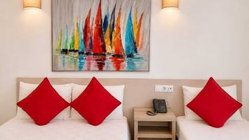 A hotel room with two beds, each with a red pillow, a bedside table with a phone, and a painting of colorful sailboats on the wall.