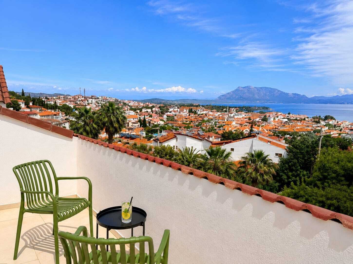 A balcony with green chairs and a small table, overlooking a coastal town with red-roofed buildings and a mountain in the distance.