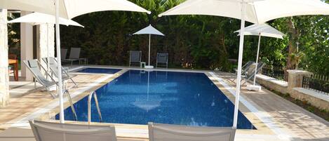 Una piscina rectangular con agua azul cristalina, rodeada de sillas de descanso y sombrillas blancas.