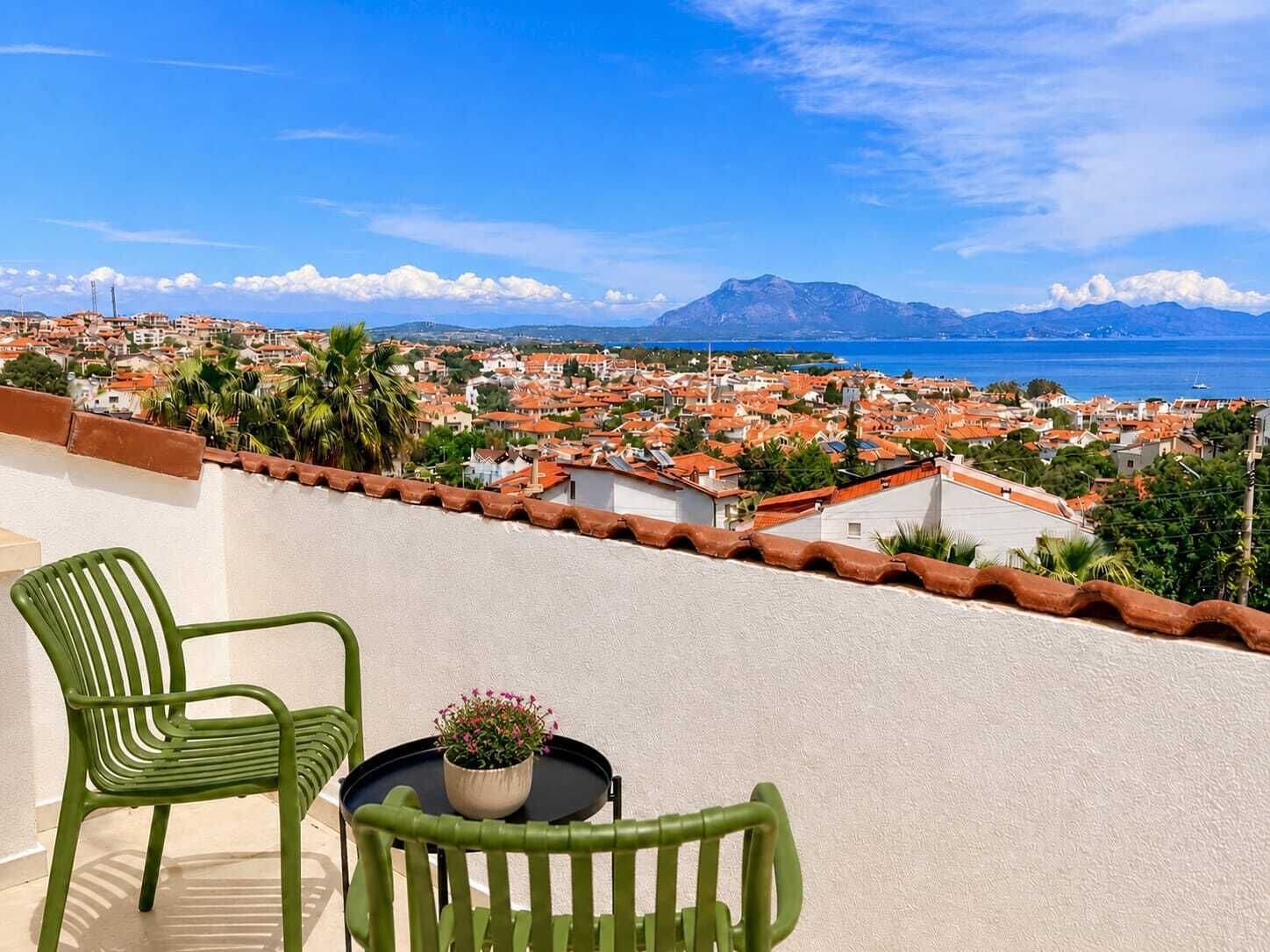 Ein Balkon mit zwei grünen Stühlen und einem Tisch mit einem Blumentopf, mit Blick auf eine Küstenstadt mit roten Dächern und einem Berg in der Ferne.