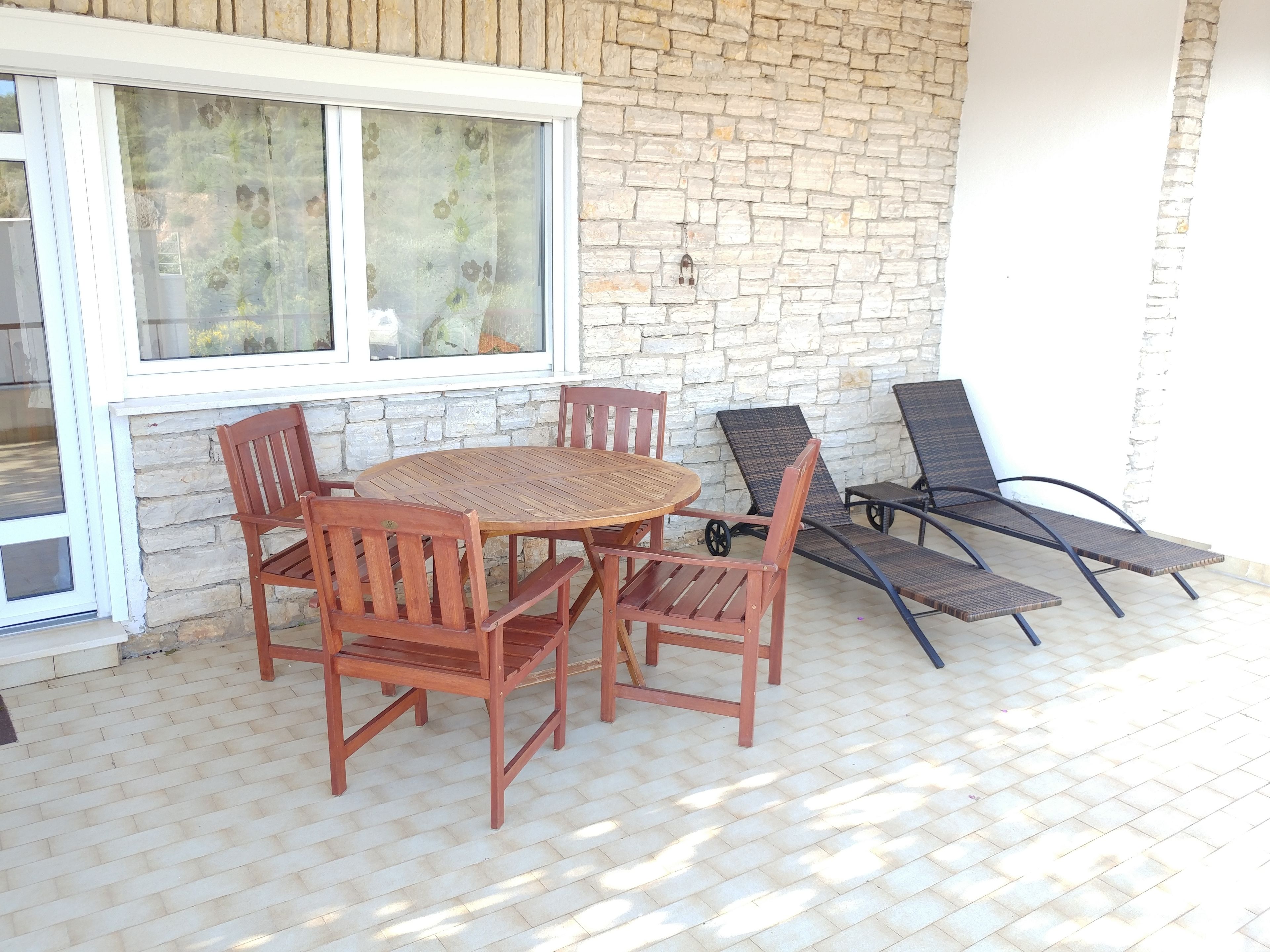 A patio with a round wooden table and chairs, a stone wall, and two lounge chairs.