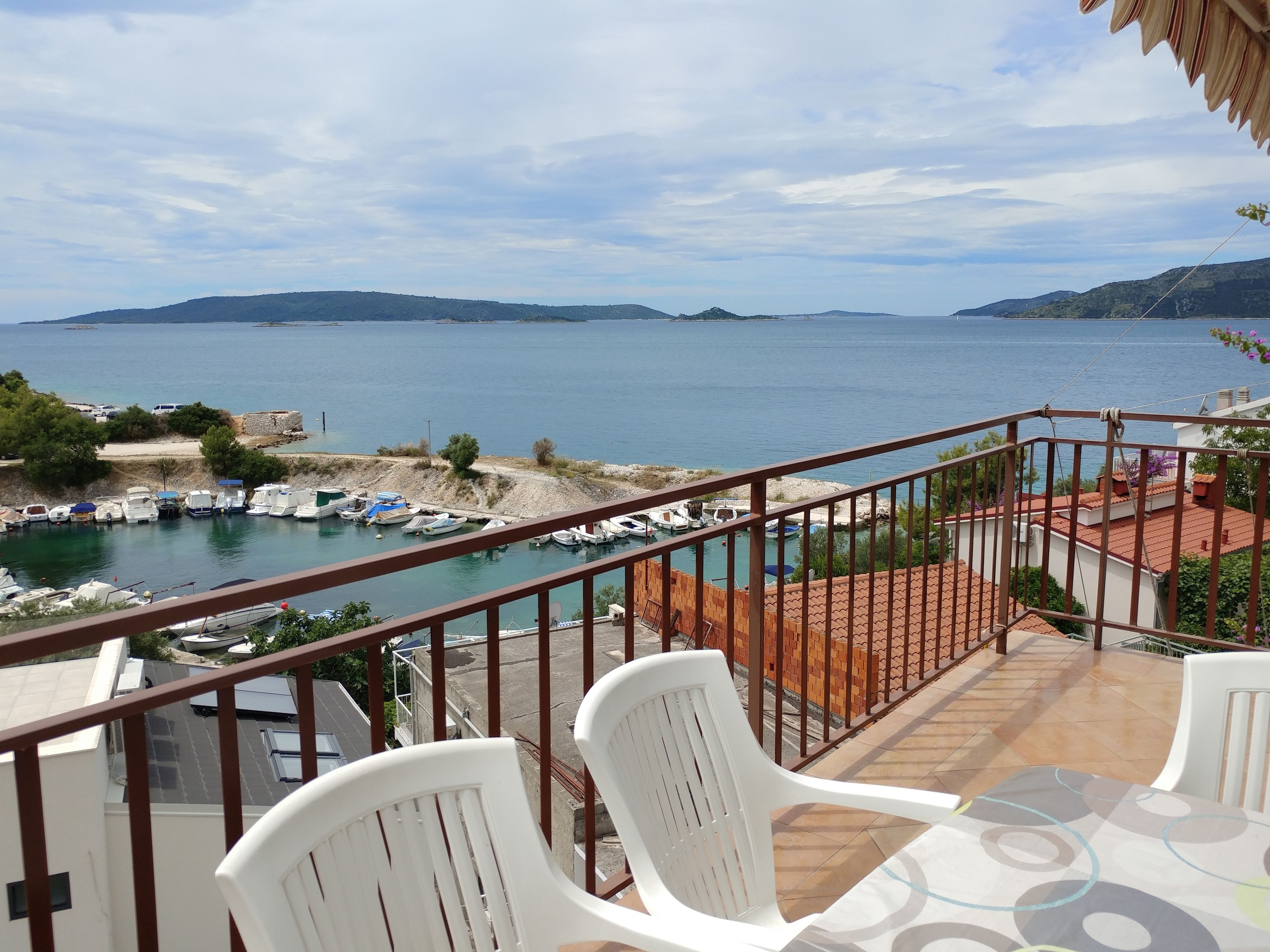 Un balcón con sillas y una mesa de plástico blanco, con vista a un puerto deportivo con barcos y una vista despejada del mar y colinas distantes.