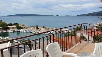 Un balcón con sillas y una mesa de plástico blanco, con vista a un puerto deportivo con barcos y una vista despejada del mar y colinas distantes.