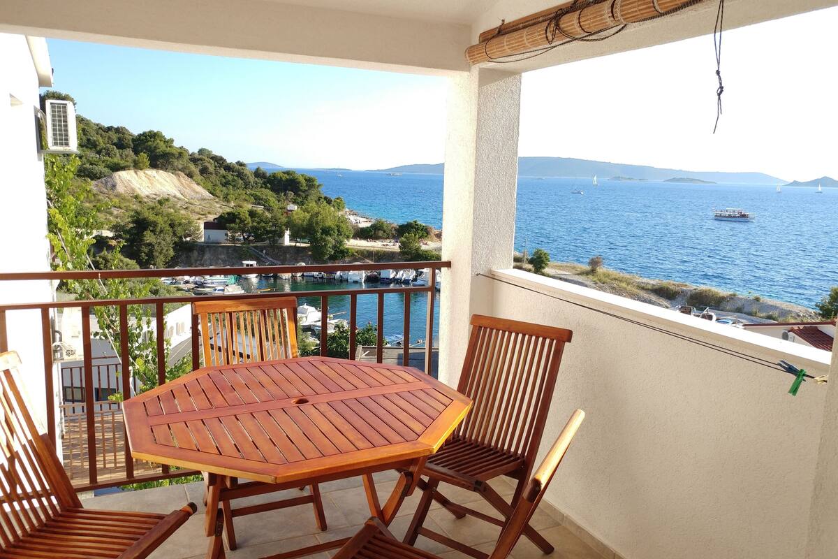 A balcony with a wooden table and chairs, overlooking a coastal area with boats and hills.