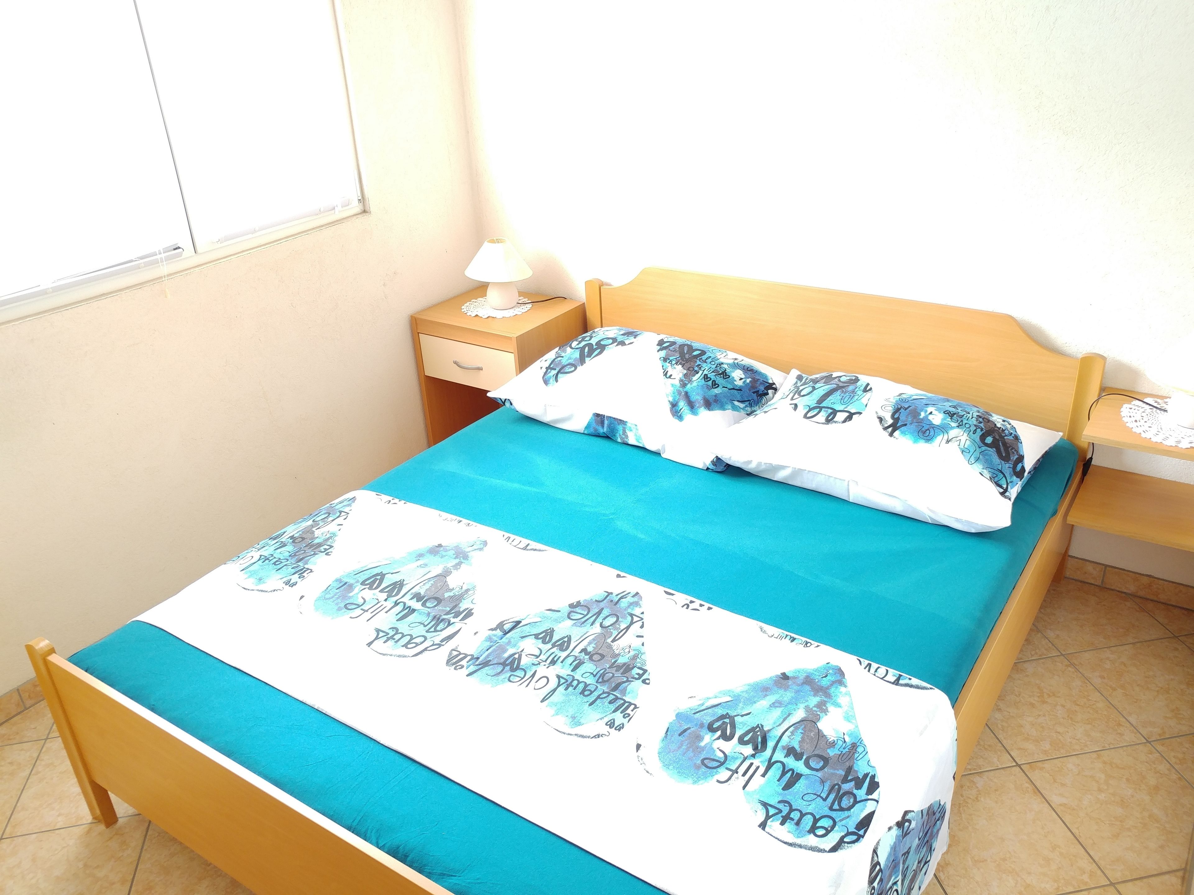 Una cama individual con una colcha azul y almohadas estampadas, una mesita de noche de madera con una lámpara, y una ventana con persianas.
