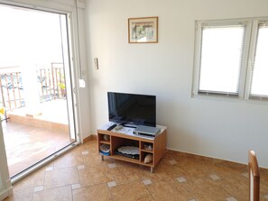 A room with a television on a wooden stand, a small table with a remote control, and a window with blinds. - Astramaris Apartments (Okrug)