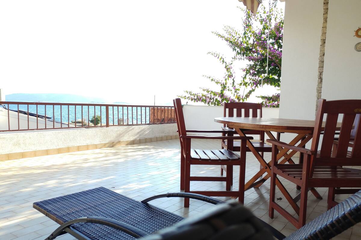A terrace with a view of the sea, furnished with wooden chairs and a table, and a wicker lounge chair.