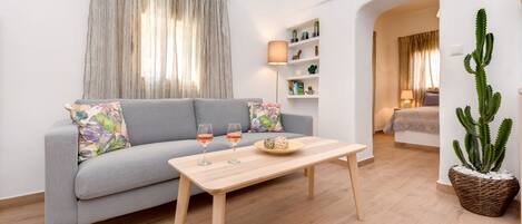 A modern living room with a grey sofa, a wooden coffee table with two glasses of wine, a cactus plant, and a bookshelf with decorative items.