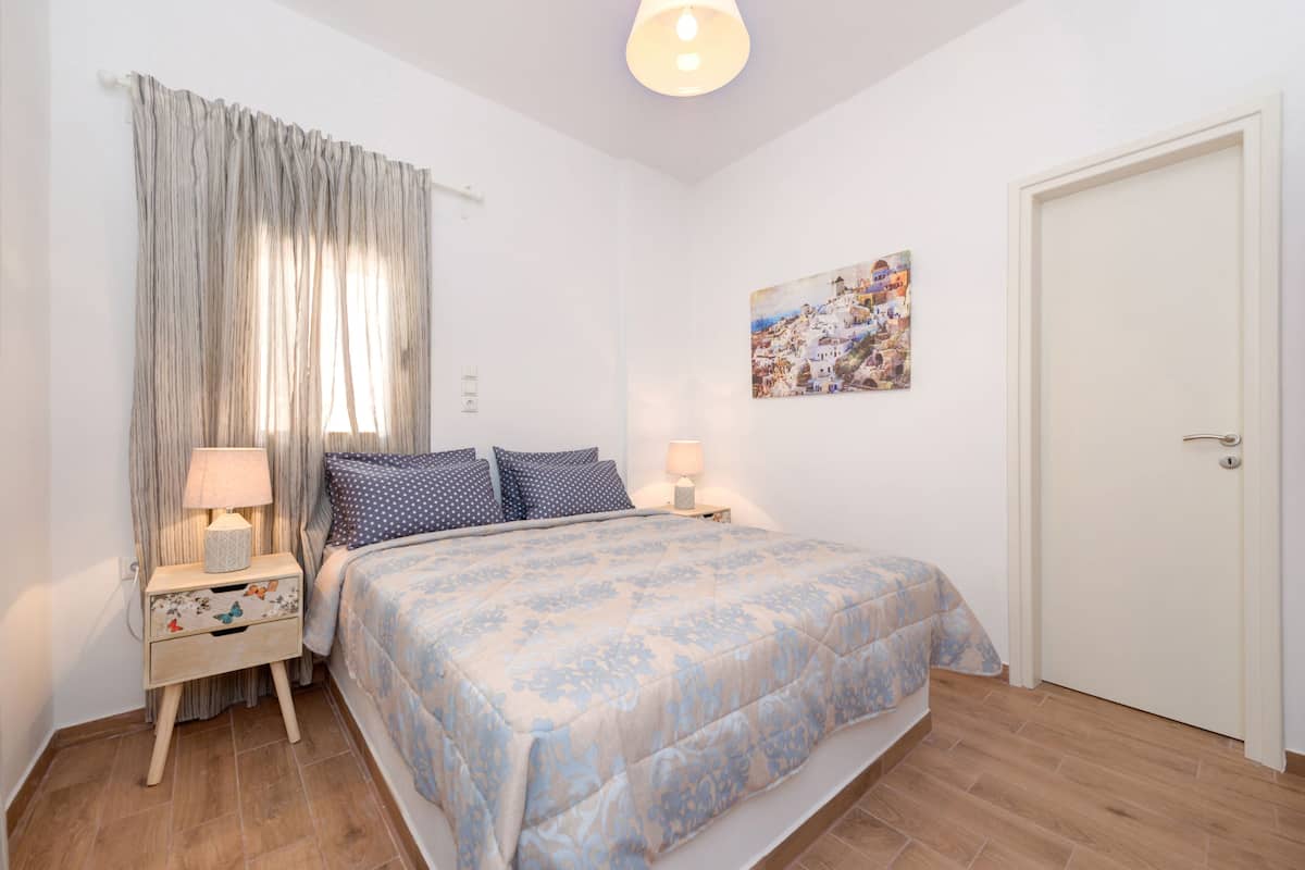 Un dormitorio con cama, dos mesitas de noche con lámparas, un ventanal con cortinas, una puerta y un cuadro colgado en la pared.