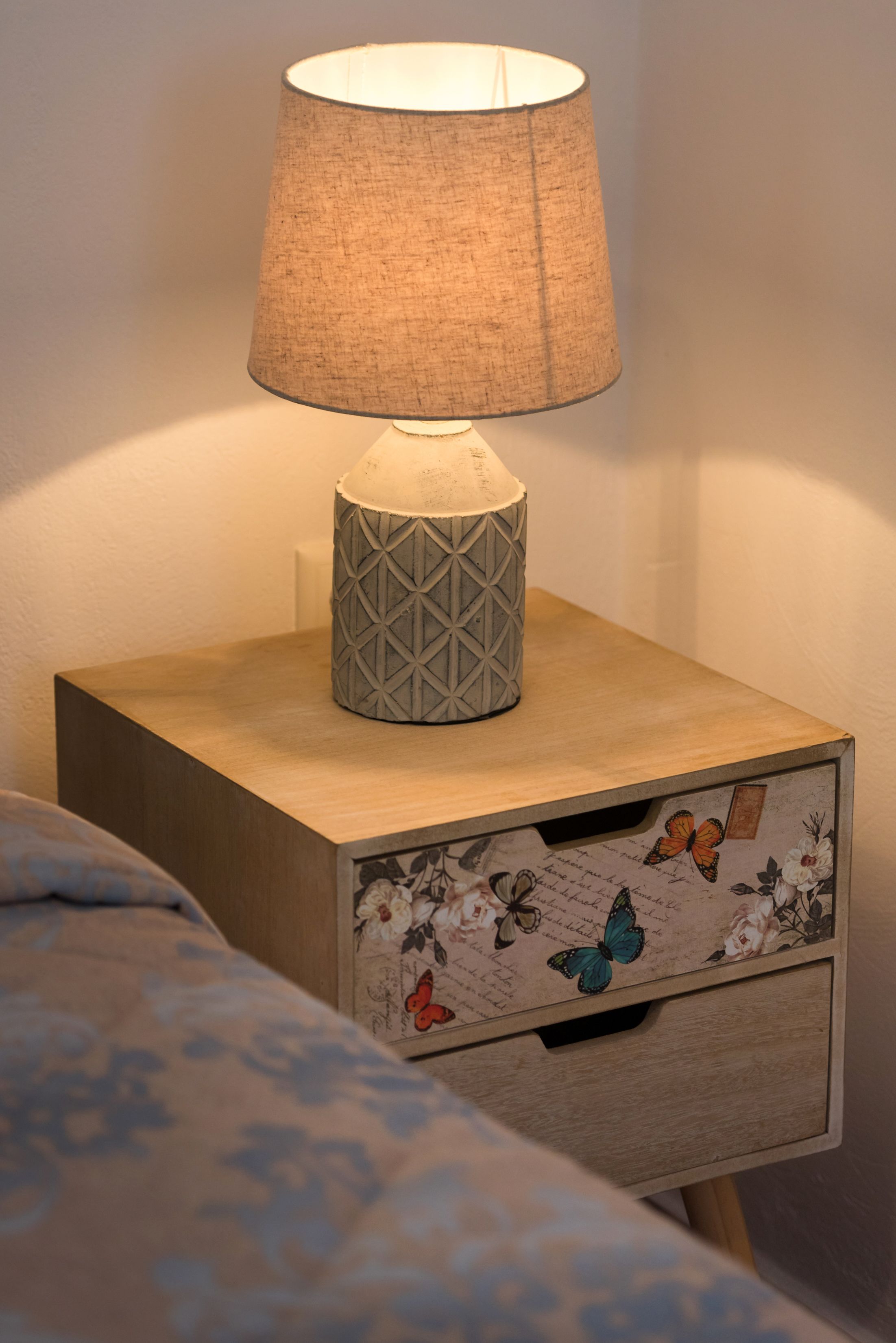 Una mesita de noche con una lámpara, un cajón estampado con motivos florales y una cama con edredón estampado.