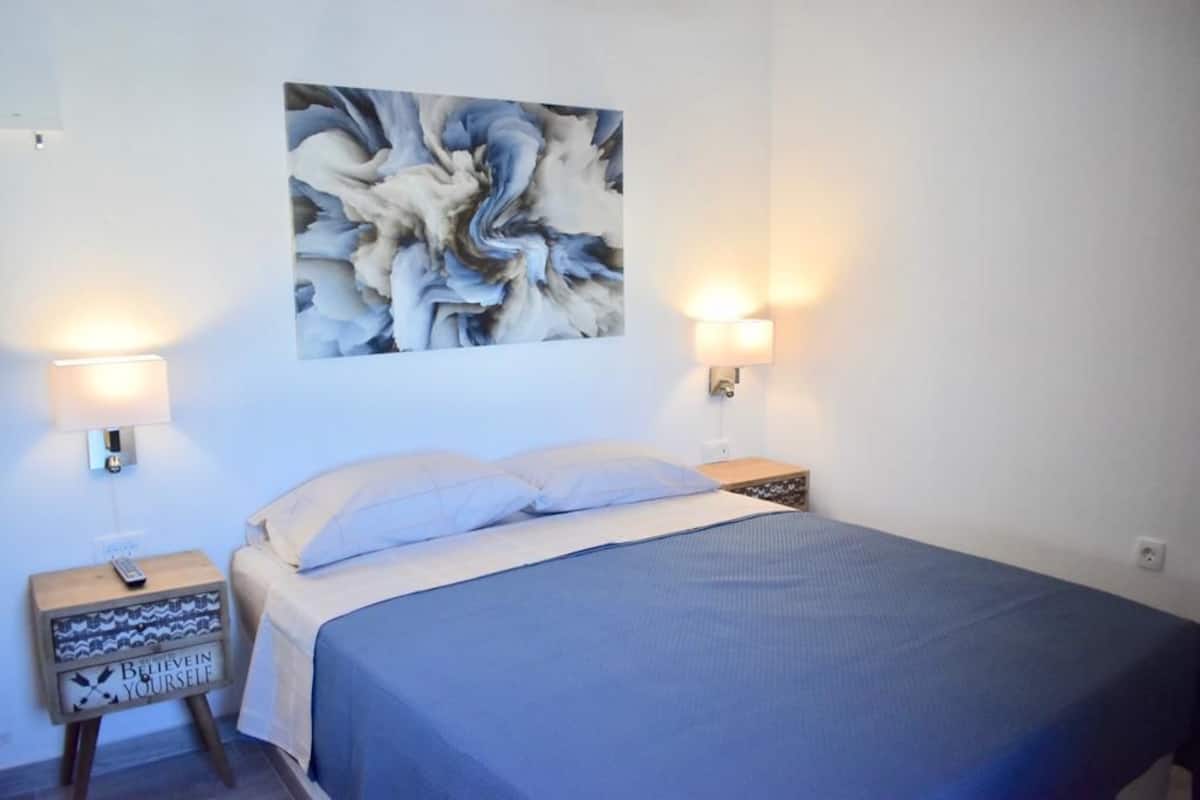 Un dormitorio con cama, dos lámparas de noche, un cuadro abstracto en la pared y una mesita de noche con un control remoto.
