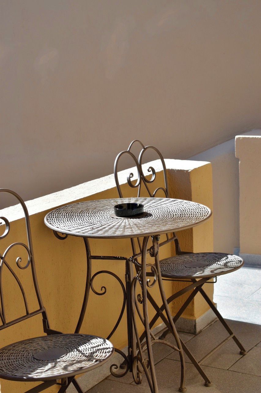 A metal table and chairs on an outdoor balcony with a beige wall and tiled floor.