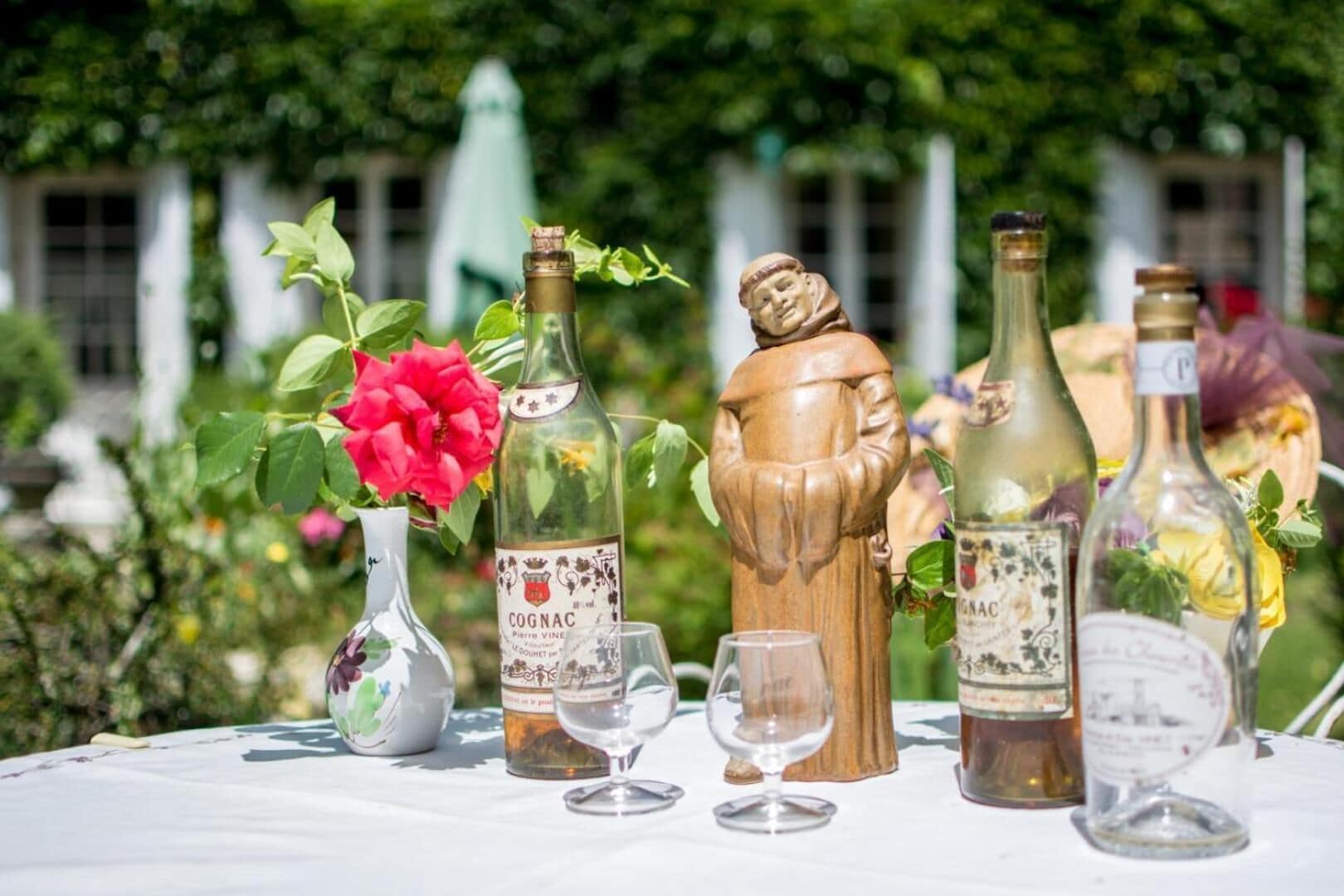 Een buitentafel gedekt met wijnflessen, glazen en een decoratief houten figuurtje.