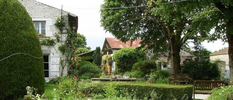 A lush garden with a variety of plants and flowers, a wooden bench, and a rustic building with a stone facade.