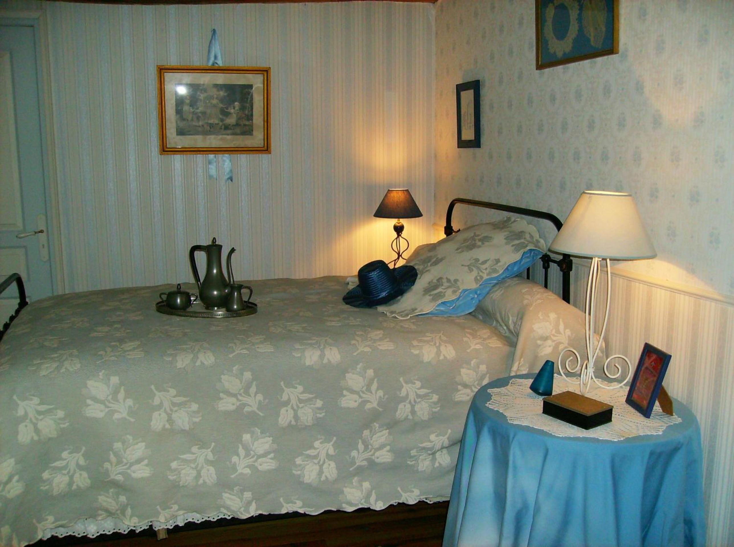 Un dormitorio con cama, una mesita de noche con lámpara, una pequeña mesa con un marco de fotos y una pared decorada con un cuadro.