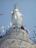 Una estatua blanca grande sobre un pedestal de piedra, con banderas y gente alrededor.