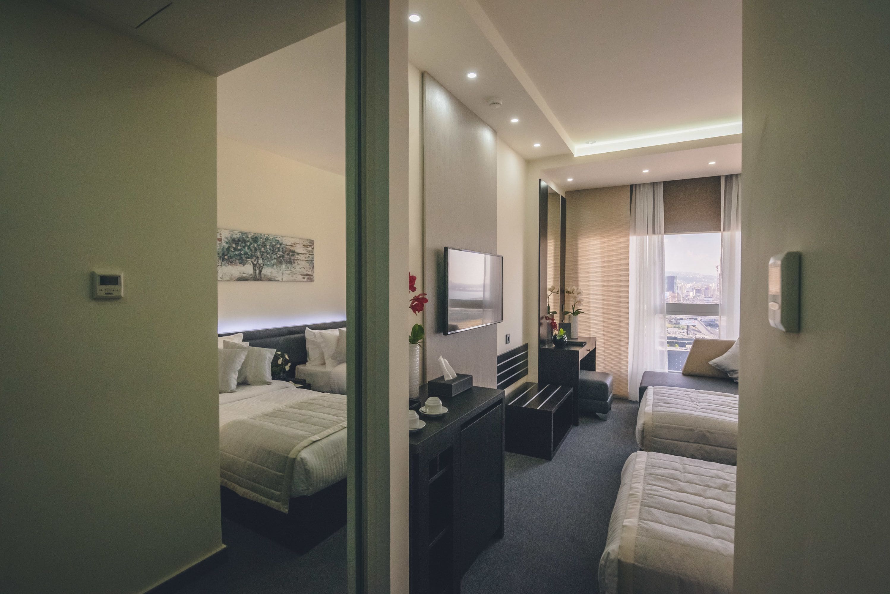 A hotel room with two beds, a desk, a flat-screen TV, a window with a city view, and a painting on the wall.