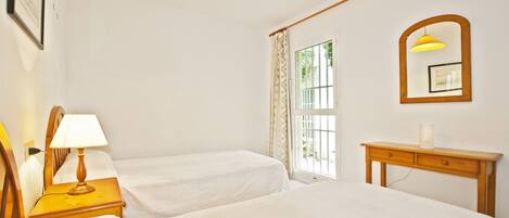 A hotel room with two beds, a bedside table with a lamp, a wooden nightstand, a mirror, and a window with curtains.