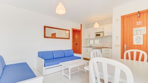 A modern living room with a blue sofa, white coffee table, and chairs. It includes a kitchen area with white cabinets and a microwave. - Apartamentos Llevant Puerto de Pollensa (Pollenca)