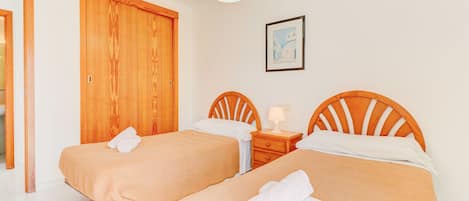 Una habitación de hotel con dos camas individuales, cabeceros de madera, una mesita de noche con lámpara, un cuadro enmarcado en la pared y un armario de madera.