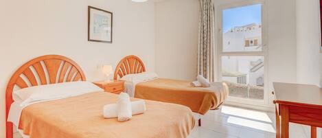A hotel room with two beds, a small table, a hanging light, a window with a view, and a framed picture on the wall.