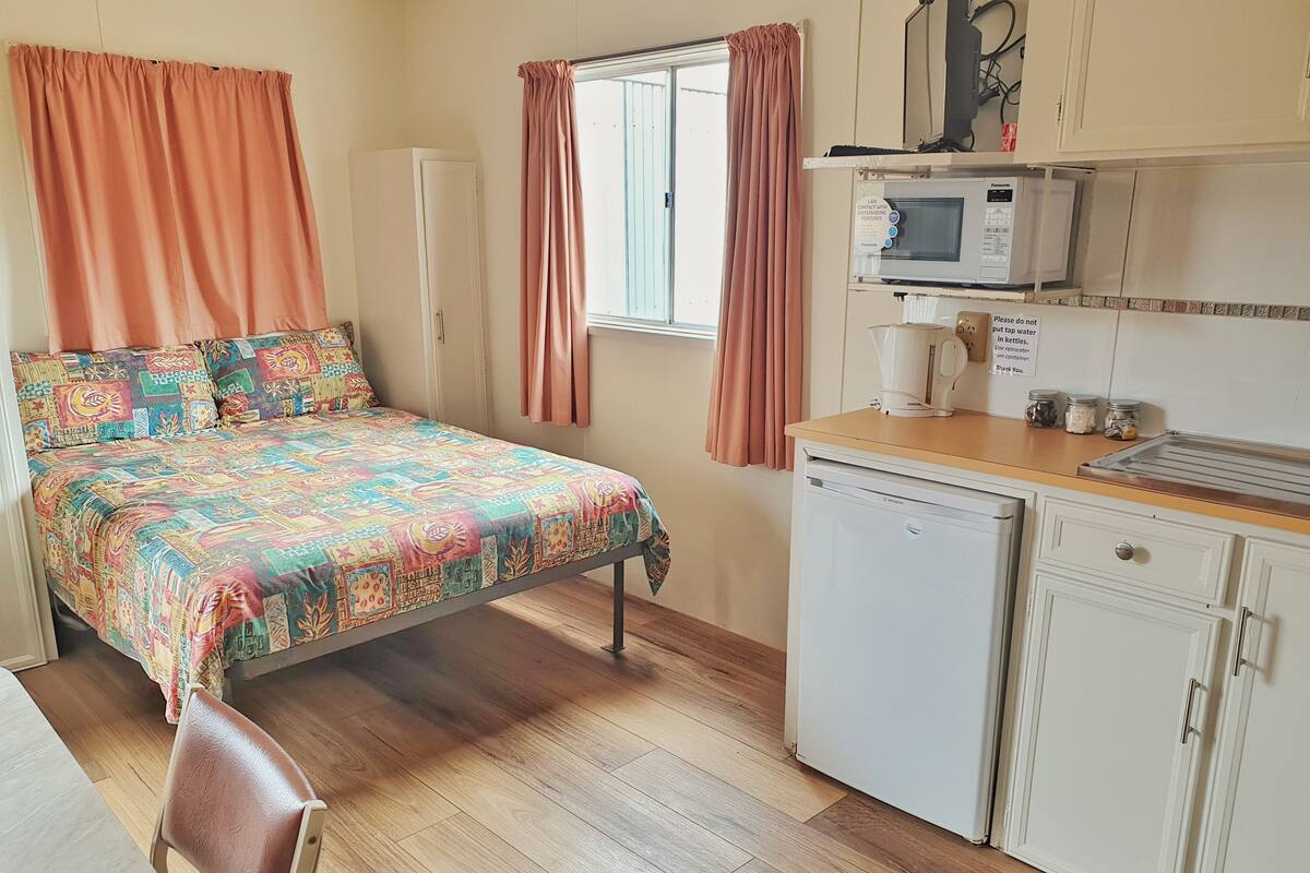 Um quarto pequeno e bem iluminado, com cama, escrivaninha, geladeira e janela com cortinas.