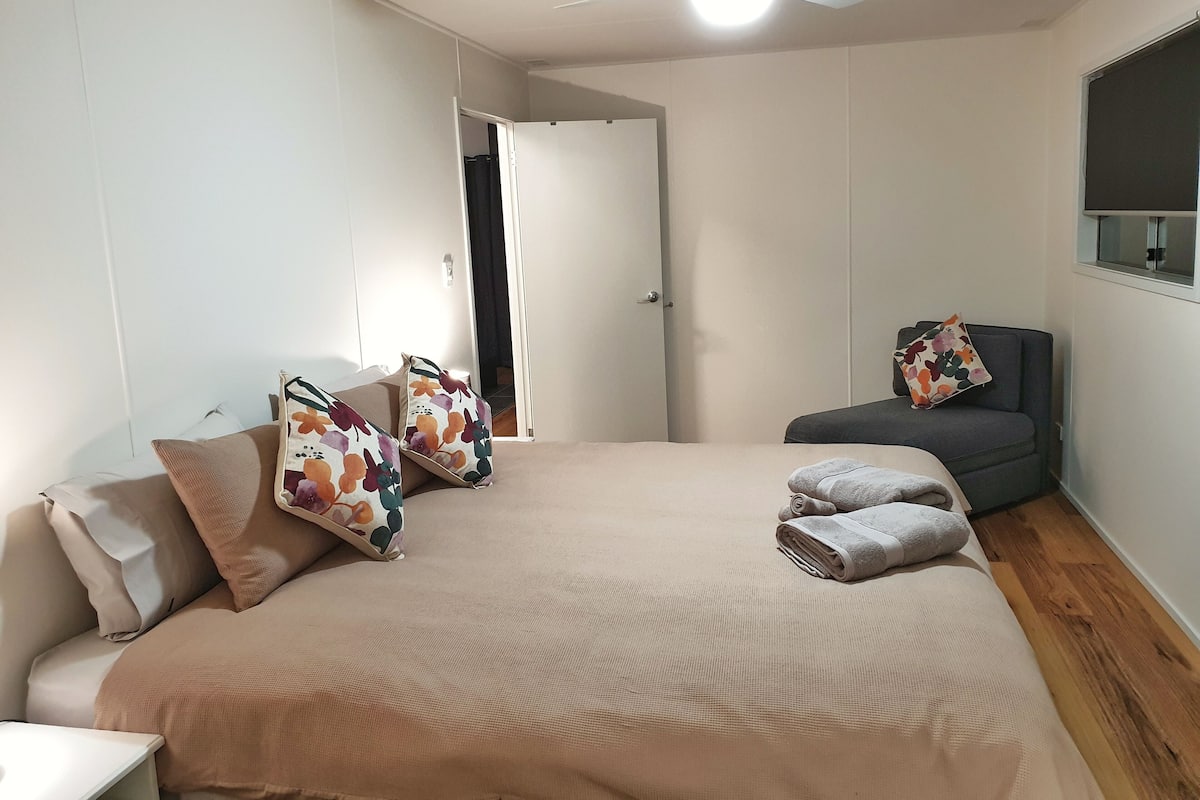 Quarto com cama, sofá cinzento, ventoinha no teto e janela com vista.