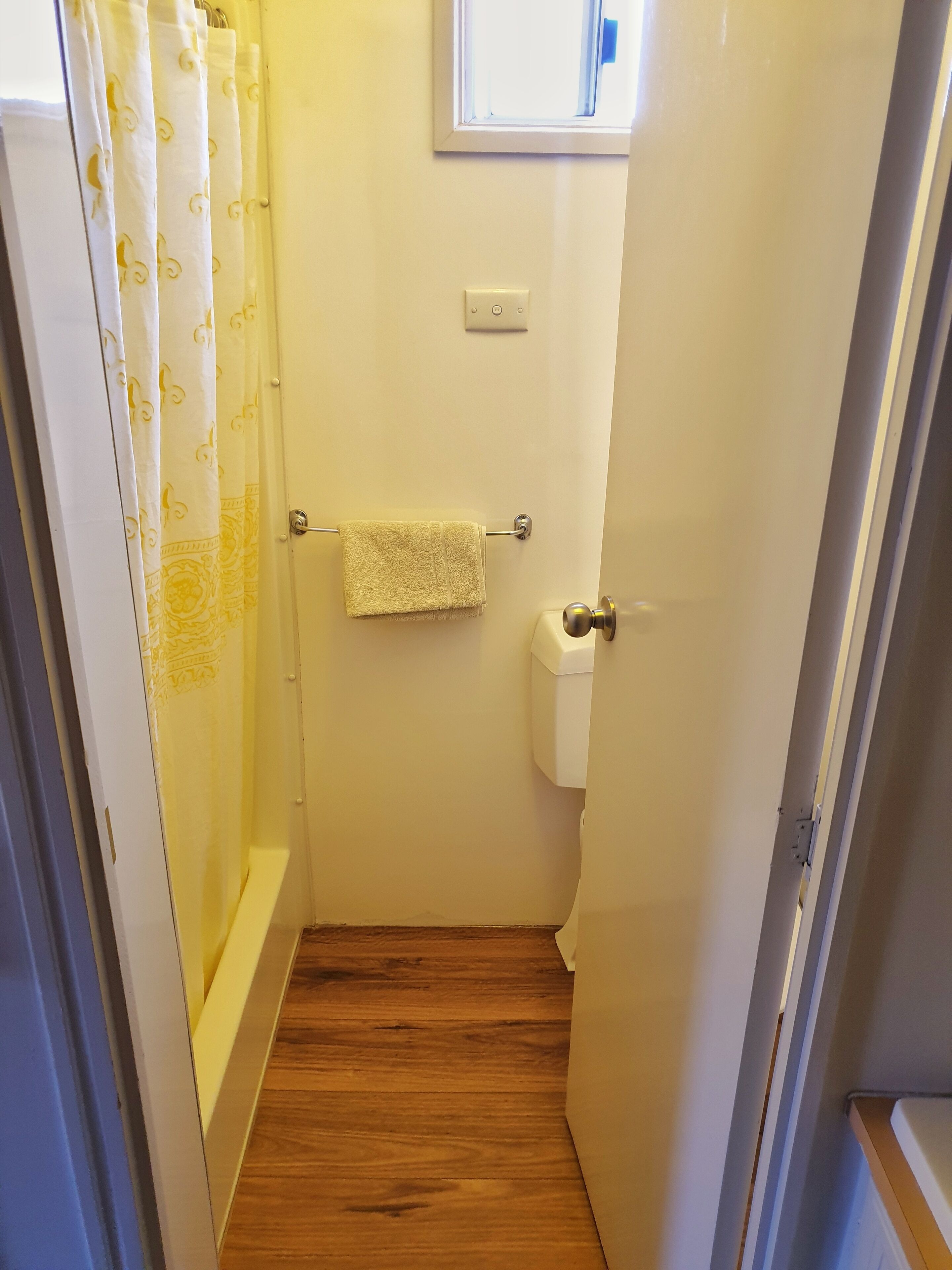 Baño pequeño con piso de madera, inodoro, una toalla colgada en un perchero y cortina de ducha estampada.