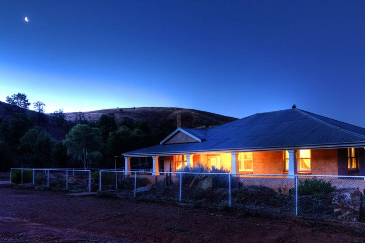 Uma casa com varanda, um quintal cercado e uma paisagem montanhosa ao fundo.