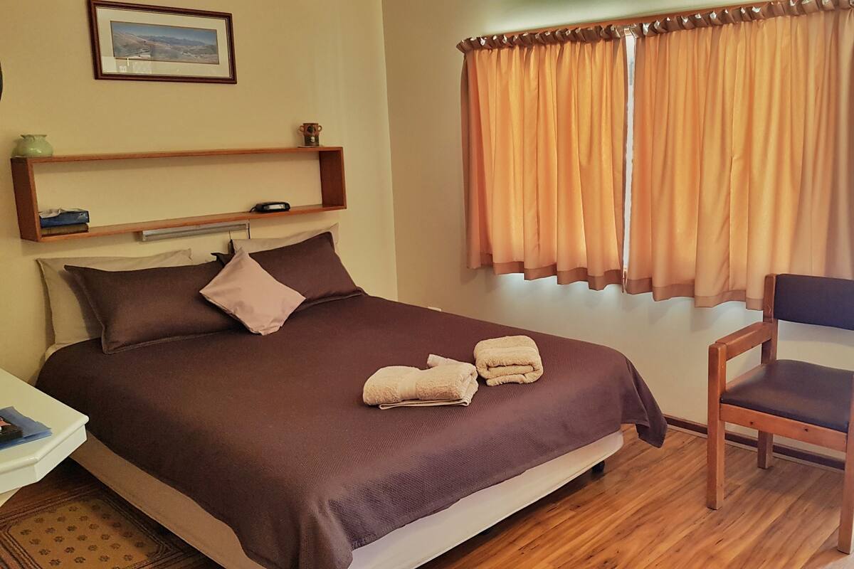 Uma cama bem arrumada com roupa de cama marrom, uma cadeira de madeira e uma prateleira com um pequeno vaso e um controle remoto.