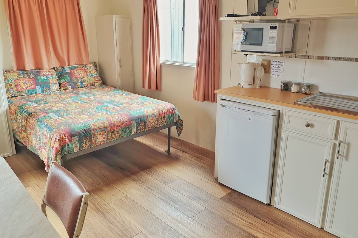 Quarto compacto com cama, pequeno espaço de cozinha, geladeira e janela com cortinas.