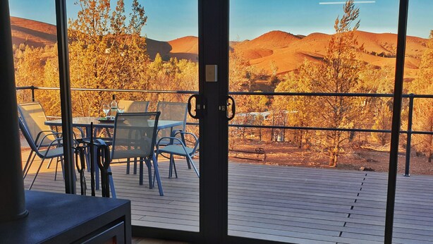 A room with a large window overlooking a scenic landscape. Inside, there is a wooden deck with a table and chairs, and a wood-burning stove. - Skytrek Willow Springs Station (Flinders Ranges)
