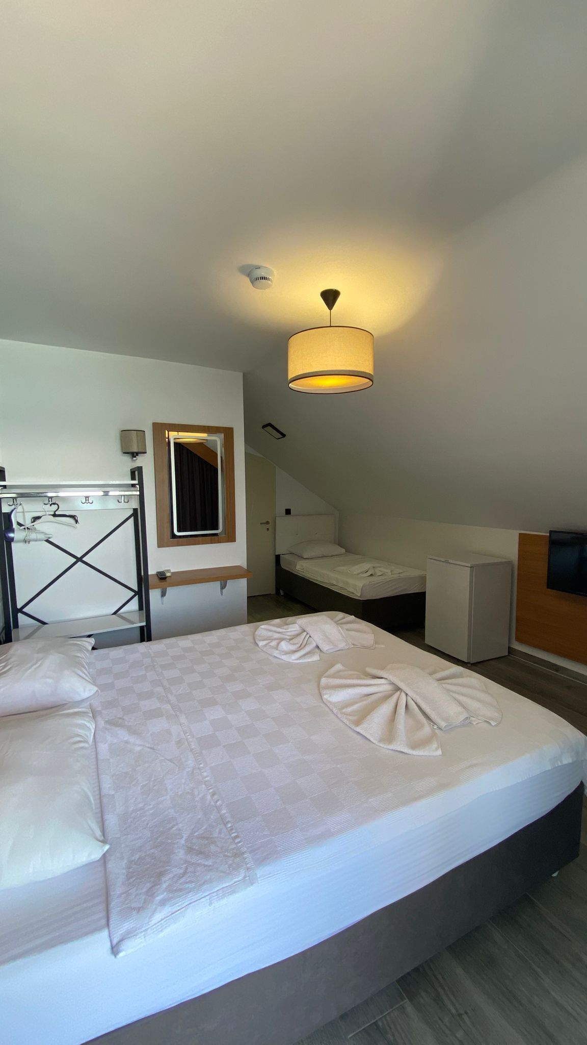 A hotel room with a bed, a desk, a mirror, a hanging light fixture, and a ceiling-mounted smoke detector.