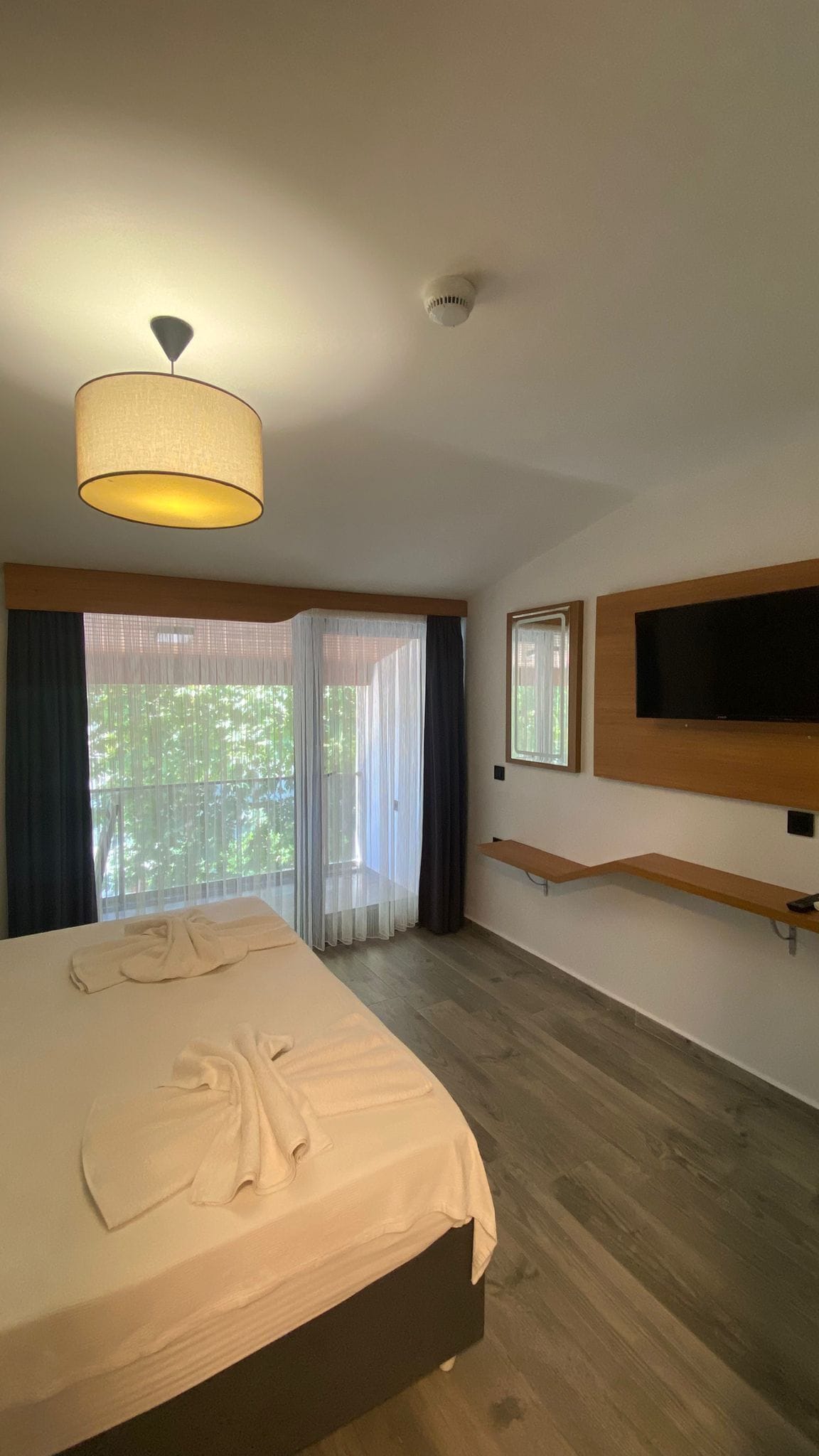 Une chambre à coucher avec un lit, un luminaire au plafond, un téléviseur fixé au mur et une fenêtre avec des rideaux.