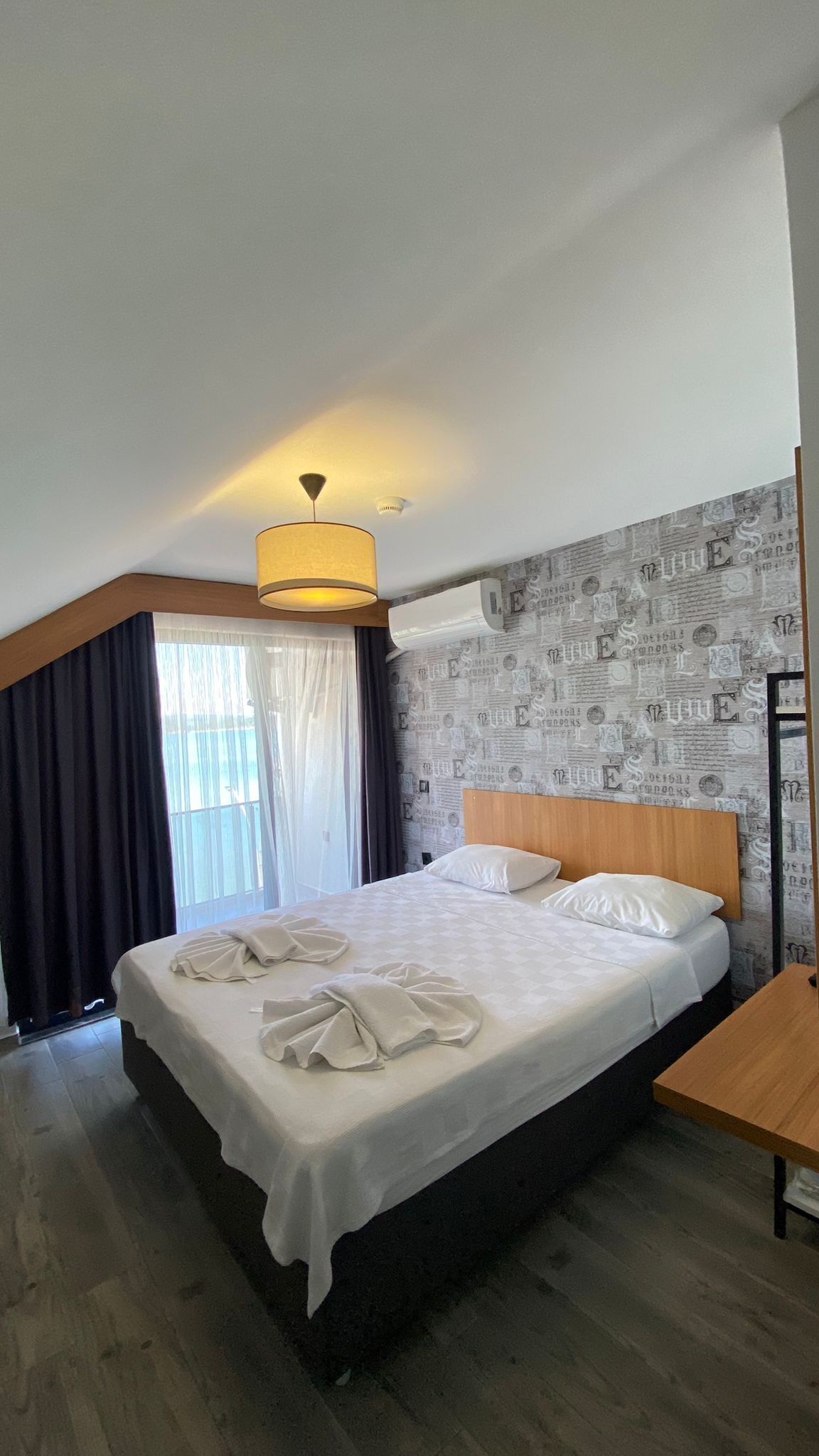 Una habitación de hotel con cama, cabecero de madera, mesita de noche y ventana con cortinas.