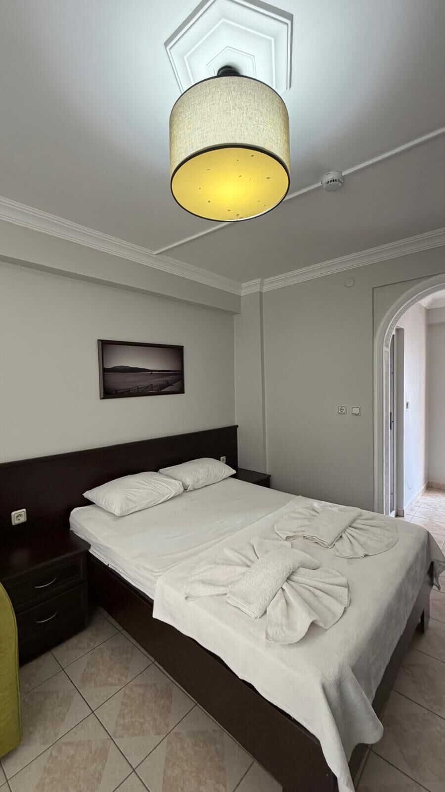 Habitación de hotel con cama, mesita de noche, lámpara de pared, un cuadro y una puerta.